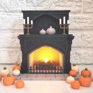 May include: A black miniature fireplace with a lit fire and two gold candle holders. White and orange pumpkins are arranged around the fireplace. The fireplace has a decorative design and is set against a white brick wall.