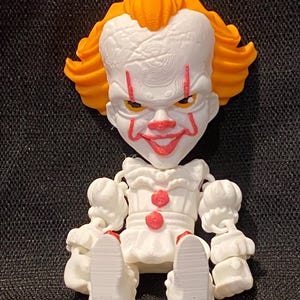 Op de afbeelding: Een 3D-geprint beeldje van een clown met een witte huid, rode lippen en oranje haar. De clown draagt een witte outfit met ruches en rode knopen. Het beeldje heeft een dreigende uitdrukking en zit.