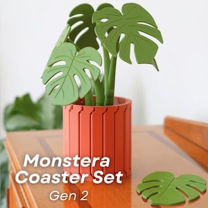 Peut inclure: Un ensemble de dessous de verre Monstera, Gen 2, avec un pot couleur terre cuite et des dessous de verre en forme de feuilles de monstera vertes. Les dessous de verre sont en forme de feuilles de monstera et sont placés sur une surface en bois. Le texte "Monstera Coaster Set Gen 2" est affiché.