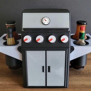 Może przedstawiać: Czarny i szary pojemnik na przyprawy w kształcie grilla z termometrem i czterema pokrętłami. Posiada dwie boczne tace z butelkami sosów i przypraw. Drzwi poniżej są szare. Zabawny dodatek do kuchni.