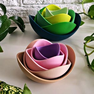 Puede incluir: Juego de cuencos anidados de colores verde, amarillo, rosa, morado y beige. Los cuencos están dispuestos en una configuración anidada, con el cuenco más grande de color verde oscuro. Los cuencos están sobre una superficie blanca con plantas verdes.