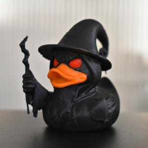 Op de afbeelding: Een zwarte rubberen eend die een zwarte heksenhoed draagt met een oranje snavel en rode ogen. De eend houdt een zwarte toverstaf in zijn linkerhand.