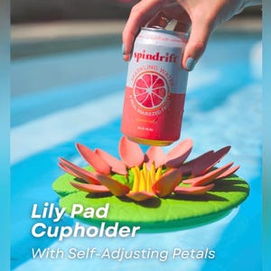 May include: A pink and white can of Spindrift Sparkling Water is held above a lily pad cupholder. The lily pad cupholder has a green base with pink petals and yellow stamens. Text on the cupholder reads "Lily Pad Cupholder With Self-Adjusting Petals."