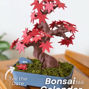 May include: A miniature bonsai tree calendar with red maple leaves and a brown trunk, set in a blue rectangular pot filled with green moss. The calendar displays "Dec 27" and the text "Bonsai 365 Calendar".