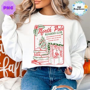 May include: White sweatshirt featuring a red and green graphic with the text "North Pole Readers Club." The design includes a Christmas tree, books, a fireplace, and a gift. The sweatshirt is a festive holiday apparel item.