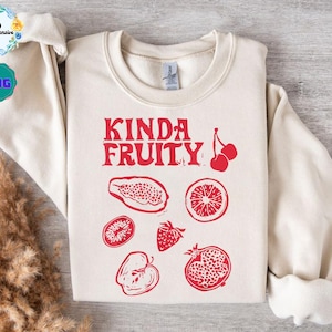 May include: Cream-colored sweatshirt with the text "Kinda Fruity" in red, along with red illustrations of various fruits. The fruits include a papaya, kiwi, orange slice, strawberry, apple, pomegranate, and cherries.