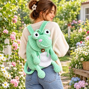 May include: A light green crochet backpack shaped like a frog with large white and black eyes, pink cheeks, a white belly patch, and a small brown button closure. The backpack features two shoulder straps and dangling frog legs and arms.