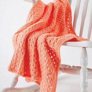 May include: A vibrant coral-colored crochet blanket featuring a repeating textured stitch pattern with small openwork gaps. The blanket is draped over the back and seat of a white wooden rocking chair with vertical slats and turned legs.