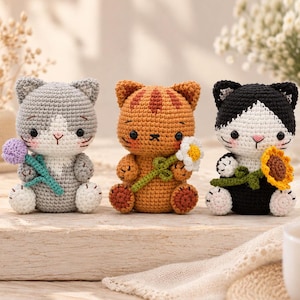 Può includere: Tre statuine di gatti all'uncinetto in fila. Il gatto a sinistra è grigio e bianco e tiene un fiore viola. Il gatto al centro è arancione a righe e tiene una margherita bianca. Il gatto a destra è nero e bianco e tiene un girasole giallo.