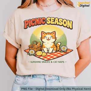 May include: Beige t-shirt featuring a cartoon cat on a picnic blanket with the words "PICNIC SEASON". The design includes a picnic basket, daisies, and the text "SUNSHINE SNACKS & CAT NAPS".