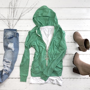 May include: A green hooded sweatshirt with a zipper and a white undershirt. The sweatshirt is on a white wooden background with a pair of blue jeans and two brown boots.