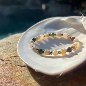 May include: A beaded bracelet with a mix of natural shell, green, and brown beads, interspersed with small gold-colored spacers. The bracelet rests on a white seashell, with a blurred background of water and stone.