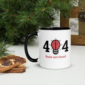 May include: White ceramic mug with a black handle and interior. The mug features the numbers "404" with a red brain inside a lightbulb graphic, and the text "Brain not found" in a red font. The mug is on a white surface.