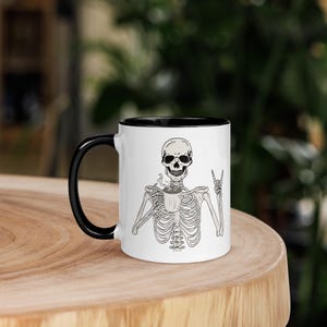 Puede incluir: Taza de café de cerámica blanca con asa e interior negros. La taza presenta una ilustración de un esqueleto sosteniendo una taza de café y haciendo un gesto de rock-on con la mano. La taza está sobre una superficie de madera.