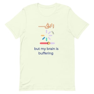 May include: A light green t-shirt featuring a colourful loading symbol and the Arabic text "...الآن". The phrase "but my brain is buffering" is printed below the symbol.