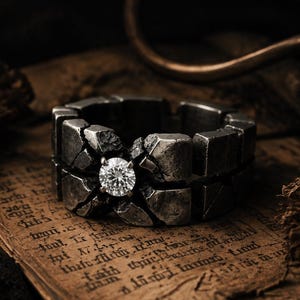 May include: A dark, weathered metal ring with a textured, cracked stone-like surface and a single, brilliant-cut clear round gemstone set prominently in the center of the band, showcasing a rugged, industrial aesthetic with deep gray tones.