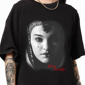 May include: Black t-shirt featuring a grayscale portrait of a woman with ornate headwear. The text "Queen Amidala" is printed in red script below the portrait. The shirt has short sleeves and a crew neck.