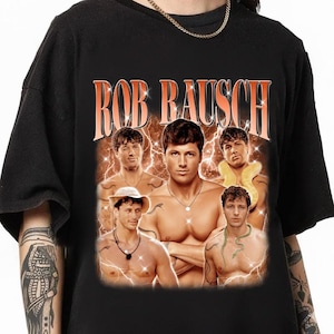 May include: Black t-shirt featuring a vintage-style graphic with the text "ROB RAUSCH" in orange. The graphic includes multiple images of a man with snakes and lightning bolt effects, creating a retro aesthetic.