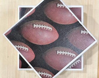 May include: Square ceramic tile with a football design. The tile features a repeating pattern of brown footballs with white laces on a black background. The tile has a white border and is set on a light wood surface.