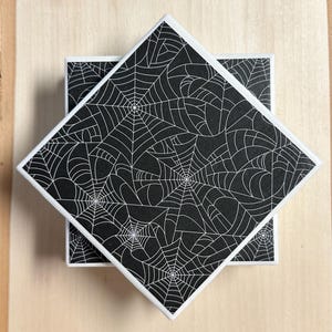 May include: Square coasters with a black background and a white spiderweb design. The coasters have white edges and are stacked on top of each other. The coasters are on a light wood surface.