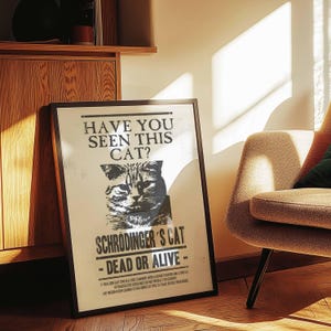 Op de afbeelding: Een ingelijste poster met een zwarte rand, met een grijswaardenafbeelding van een kat en de tekst "HAVE YOU SEEN THIS CAT?" en "SCHRODINGER'S CAT - DEAD OR ALIVE -." De poster leunt tegen een muur in de buurt van een houten kast en een beige fauteuil.