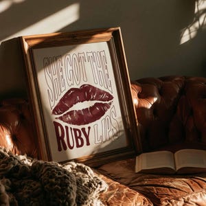 May include: A framed art print featuring the text "SHE GOT THE RUBY LIPS" and a burgundy lip illustration. The print is on a brown leather sofa with a blanket and an open book.