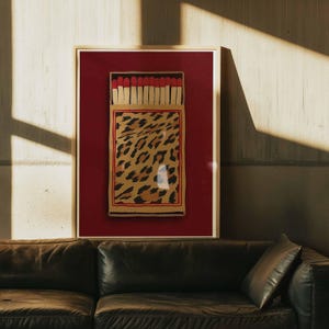 Puede incluir: Obra de arte enmarcada que presenta un diseño de caja de cerillas con un estampado de leopardo. Las cerillas tienen puntas rojas y la caja está sobre un fondo rojo. La obra de arte está en un marco de madera y se exhibe sobre un sofá de cuero negro.