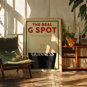 Op de afbeelding: Een ingelijste poster met de tekst "THE REAL G SPOT" in rode letters. Daaronder een afbeelding van een Guinness pintglas met het Guinness logo. De poster staat in een kamer met een houten stoel en een boekenrek.