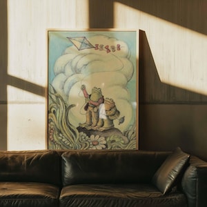 May include: A framed illustration of two frog characters, one flying a kite. The artwork uses soft colours, including green, brown, and blue. The frame is light wood, and the artwork is displayed above a dark brown leather sofa. The illustration has a whimsical style.