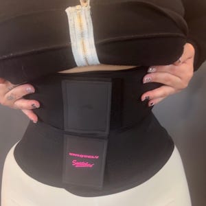 Puede incluir: Un cinturón de cintura negro con un panel frontal rectangular. El panel tiene las palabras "Uniquely Snatched" en escritura rosa. El cinturón se usa sobre pantalones blancos y debajo de una chaqueta negra con cremallera.