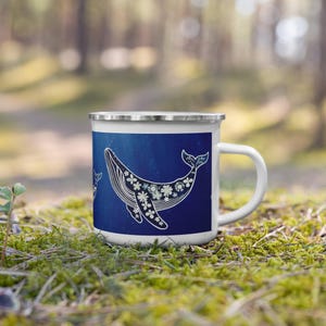May include: White enamel mug with a blue background and a whale design. The whale is adorned with white and light blue floral patterns. The mug has a silver rim and a white handle. The mug is on green moss.