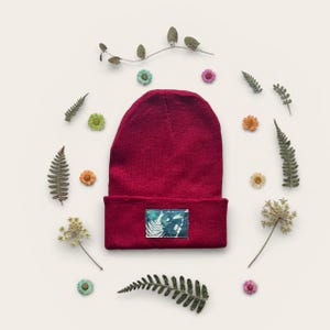 Puede incluir: Un gorro rojo oscuro con un parche rectangular con un diseño de helecho. El gorro está rodeado de flores secas y helechos sobre un fondo crema. Un accesorio elegante.