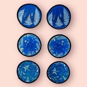 Może przedstawiać: Sześć okrągłych naszywek z niebieskim tłem i białymi wzorami paproci. Każda naszywka ma czarną obwódkę. Naszywki są ułożone w dwóch rzędach po trzy na jasnoróżowym tle.
