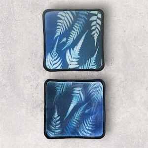 May include: Two square coasters featuring a dark blue background with a repeating light blue fern leaf pattern. Each coaster has rounded corners and a thick black stitched border, providing a consistent look for both pieces in the set.