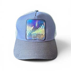 May include: A slate blue trucker hat featuring a square patch on the front panel. The patch displays a vibrant, iridescent, multi-colored background with a graphic print of two fern leaves in a light, contrasting color.