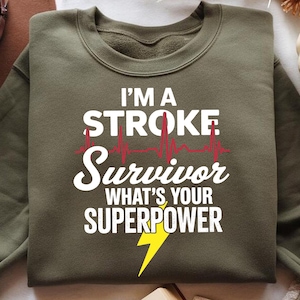 Peut inclure: Sweat-shirt vert olive avec l'inscription blanche "I'M A STROKE Survivor WHAT'S YOUR SUPERPOWER", une ligne de battement de cœur rouge et un éclair jaune. Le sweat-shirt est de style col rond.