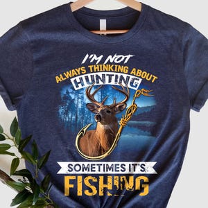 Könnte beinhalten: Ein marineblaues T-Shirt mit einer abgenutzten gelben Grafik, auf der "Ich denke nicht immer an die Jagd, manchmal ist es Angeln" steht. Die Grafik zeigt ein Reh mit Geweih und einem Angelhaken.