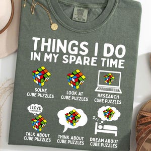 Puede incluir: Una camiseta verde con el texto "Things I do in my spare time" y una serie de ilustraciones de un cubo de Rubik. Las ilustraciones muestran a una persona resolviendo un cubo, mirando un cubo, investigando cubos, hablando de cubos, pensando en cubos y soñando con cubos.