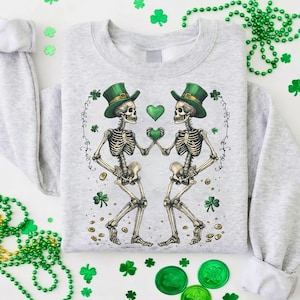 Op de afbeelding: Een lichtgrijs sweatshirt met een afbeelding van twee skeletten met groene hoge hoeden die een groen hart vasthouden. Het ontwerp bevat klavers en gouden munten. De sweatshirt heeft lange mouwen en een ronde hals.