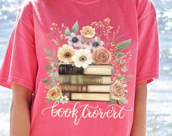 May include: A coral-colored t-shirt featuring a floral design with a stack of books. The design includes roses, anemones, and other flowers in shades of cream, tan, and pink. The words "booktrovert" are written in a cursive font below the books.