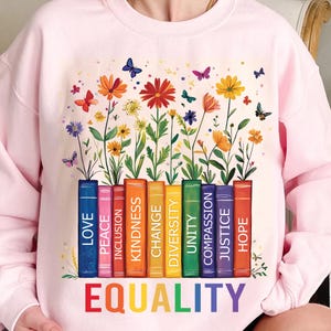 May include: Pink sweatshirt featuring a colourful floral design and a row of books with words such as "Love", "Peace", "Inclusion", "Kindness", "Change", "Diversity", "Unity", "Compassion", "Justice", and "Hope". The words are stacked on top of each other and the word "EQUALITY" is written in rainbow colours below the books.