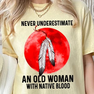 May include: A yellow t-shirt with the text "Never underestimate an old woman with native blood." The shirt features a red circle with two white feathers and a gold design.