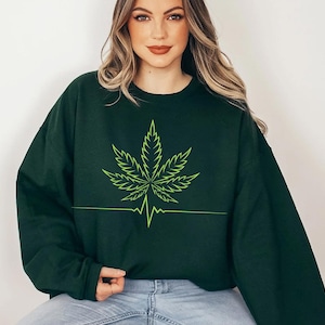 May include: Dark green sweatshirt with a neon green cannabis leaf design and a heartbeat line. The sweatshirt is a solid dark green colour and has a crew neck.