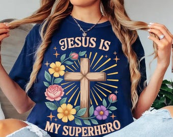 May include: Navy blue t-shirt with the text "JESUS IS MY SUPERHERO". The design features a wooden cross, surrounded by yellow, pink, and purple flowers, with sunburst rays. The shirt is a comfortable, casual wear.