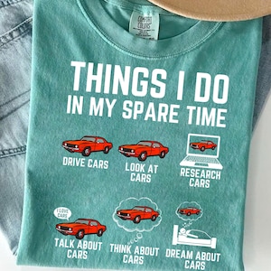 May include: A teal blue t-shirt with the text "Things I do in my spare time" and a list of activities related to cars. The activities include driving, looking at, researching, talking about, thinking about, and dreaming about cars. Each activity is illustrated with a red car.