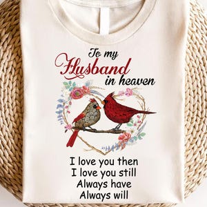 May include: A white t-shirt with a design of two red cardinals perched on a branch with a heart-shaped floral wreath. The text "To my Husband in heaven I love you then I love you still Always have Always will" is printed on the shirt.