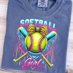 Pode incluir: Uma camiseta azul com um gráfico de uma bola de softball, dois bastões cruzados e o texto "Softball Girl". A bola de softball é amarela com costuras rosa e os bastões são azuis e dourados.