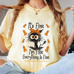 Può includere: T-shirt giallo chiaro con un gatto nero con un fiocco arancione e la scritta "It's Fine, I'm Fine, Everything Is Fine". Il design include nastri arancioni ed è destinato a sensibilizzare.