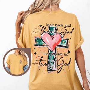 Puede incluir: Camiseta de color amarillo mostaza con un dise&ntilde;o de cruz y coraz&oacute;n. La cruz es verde con un coraz&oacute;n rosa en el centro. El texto dice "look back and thank God, look forward and trust God."