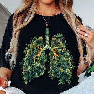 May include: Black t-shirt with a graphic of lungs made from green cannabis leaves and buds, and a brown trachea. The design is centrally placed. This is a casual, everyday wear item.